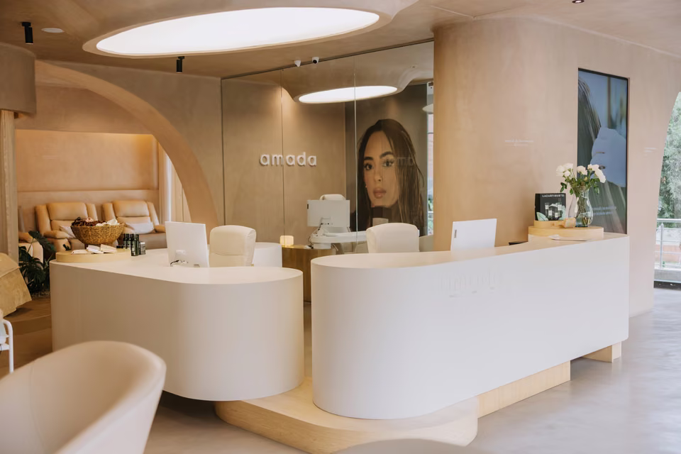 Recepción con mostradores curvos blancos bajo un arco de madera clara y el logo «amada» en la pared