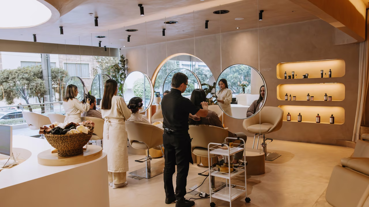 Salón de tratamientos capilares con espejos redondos, sillones beige y repisas iluminadas, donde varias especialistas atienden a clientas