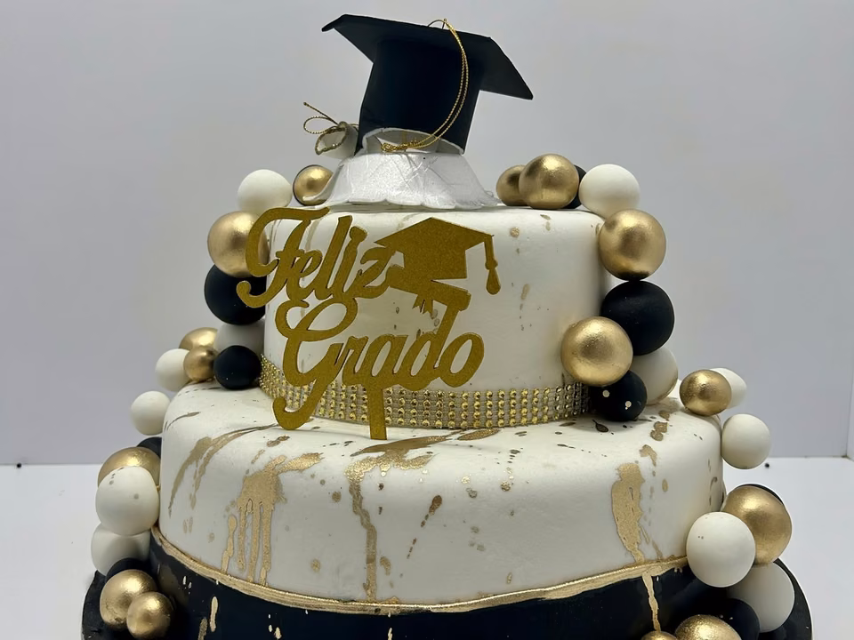 Torta de grado de dos pisos, blanca con chorreado dorado, esferas negras y doradas, birrete y letrero «Feliz Grado»