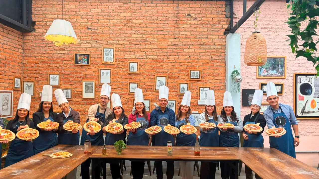 Corporate team wearing chef hats and aprons, each person holding a pizza, at a brick-walled cooking workshop in Bogotá